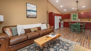 Una sala de estar con un sofá de cuero marrón, una mesa de centro de madera y un tapete estampado. Se ve una cocina con armarios rojos y un comedor con sillas verdes al fondo.