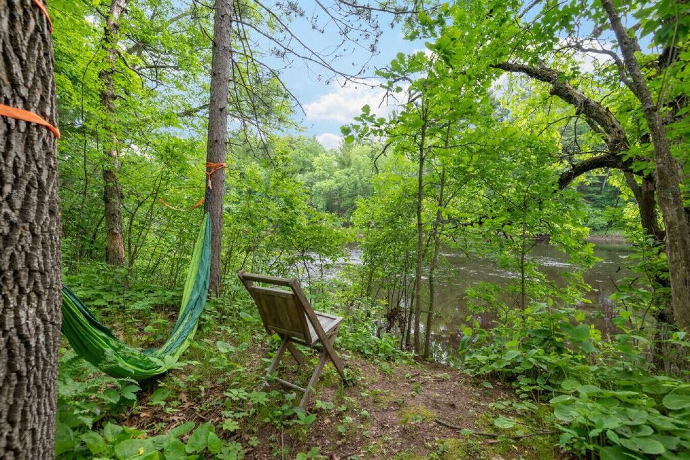 A hammock tied to a tree, a wooden chair, and a river in a forested area.
