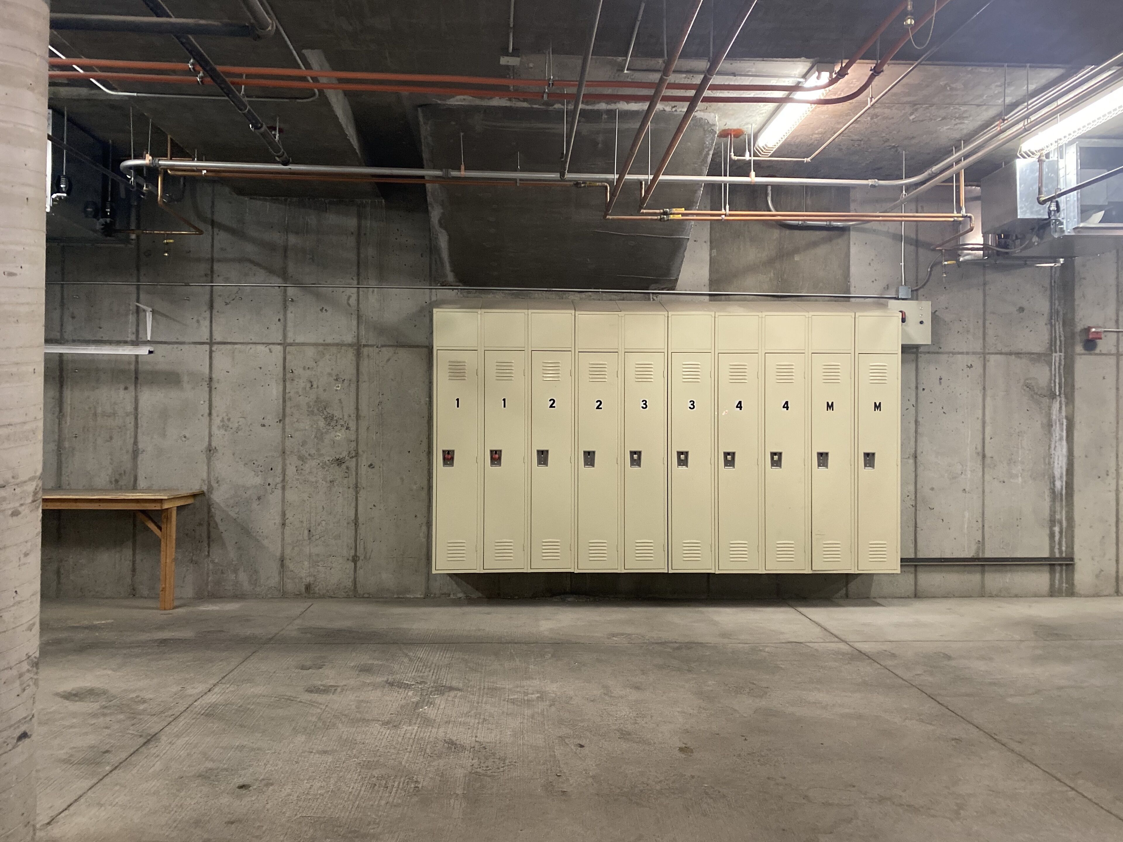 A locker room with beige lockers numbered 1 to 4 and M, concrete walls, and exposed pipes on the ceiling.