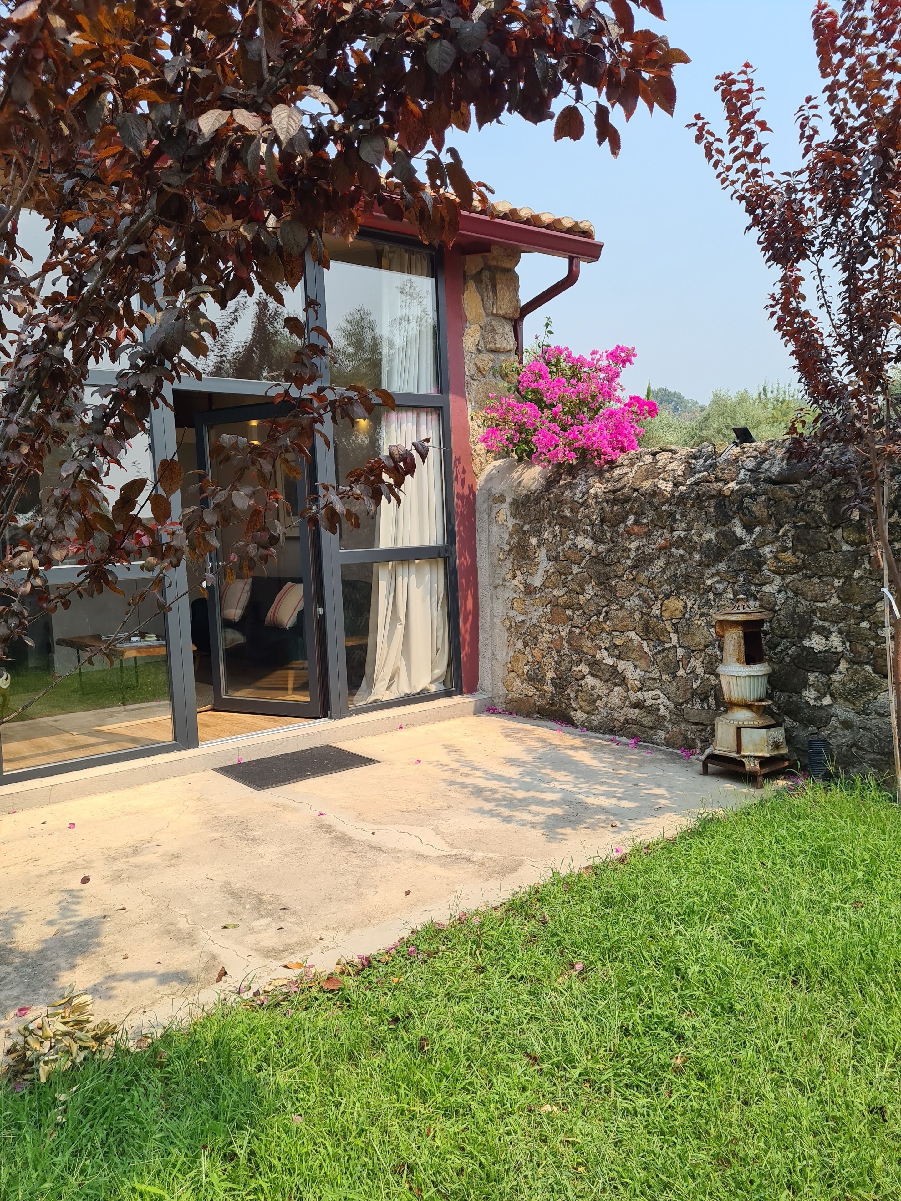 A modern house with a stone wall, a glass door, and a small garden area with grass and pink flowers.