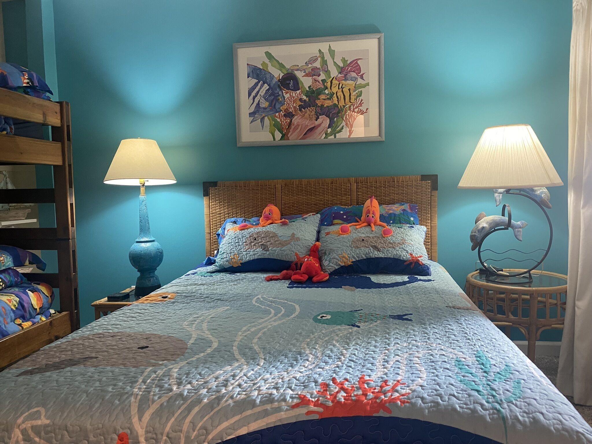 A bed with a blue ocean-themed bedspread, two pillows, and a framed fish painting above it. There are two lamps on either side of the bed.
