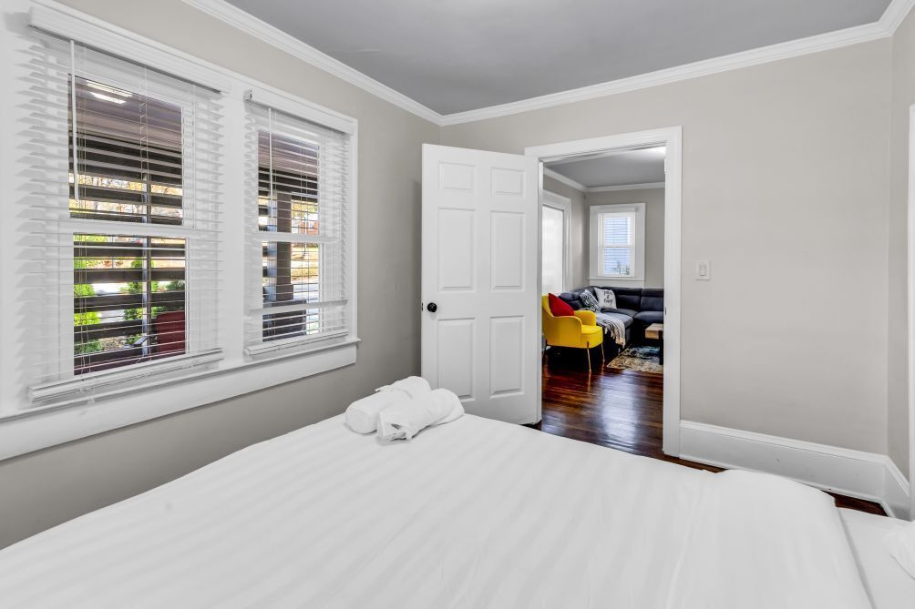A bedroom with a bed, white curtains, a door leading to another room, and a view of a living area with a sofa and a chair.