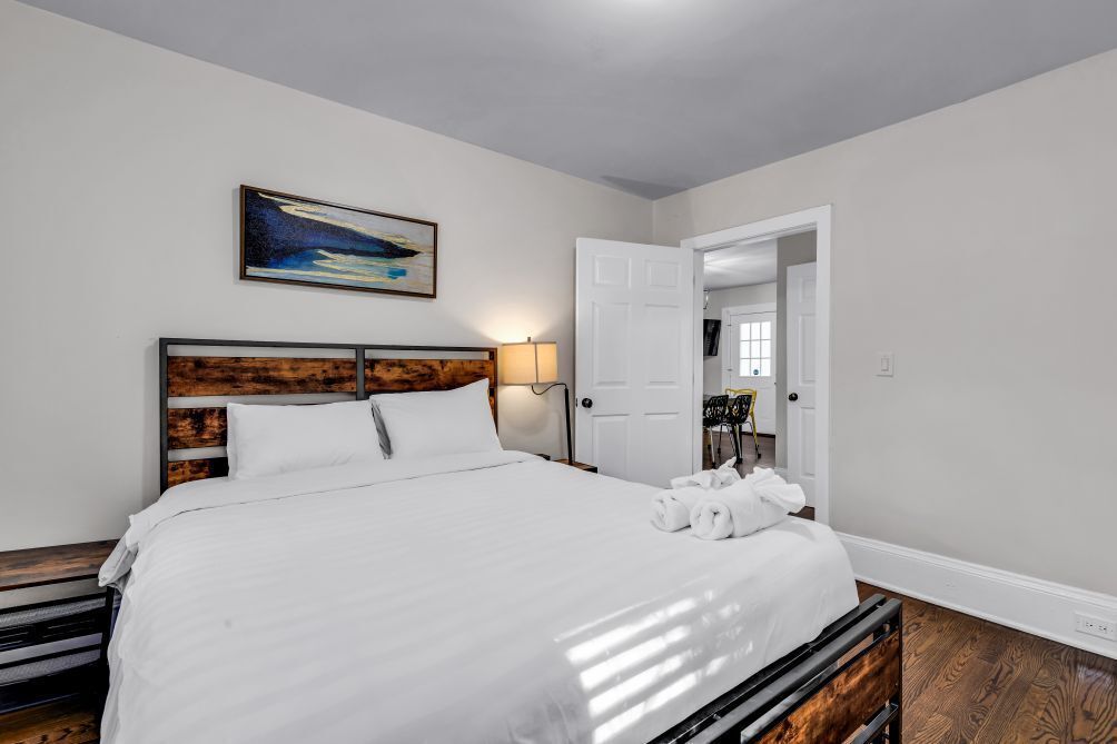 A neatly made bed with white linens, a wooden headboard, and a painting above it.
