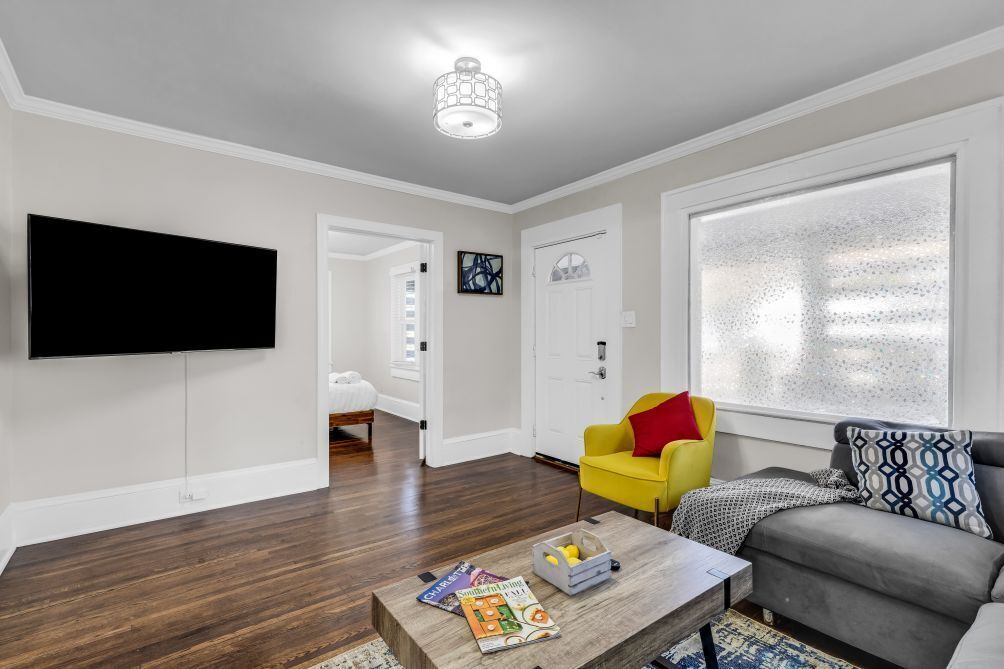 A modern living room with a flat-screen TV, a yellow armchair, a grey sofa, and a coffee table with magazines.