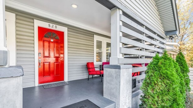 A modern house with a red door marked "820", a covered porch with red chairs, and a small green bush.