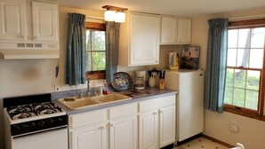 A kitchen with white cabinets, a stove, a sink, and a refrigerator. There is a window with curtains, a rug, and a chair.