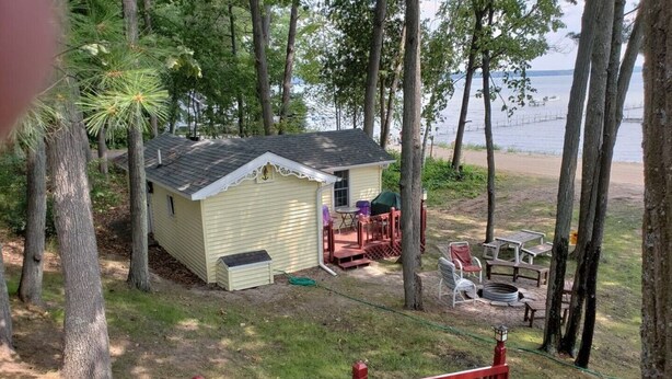 A small, single-story cabin with a covered patio area, surrounded by trees and a view of a body of water.