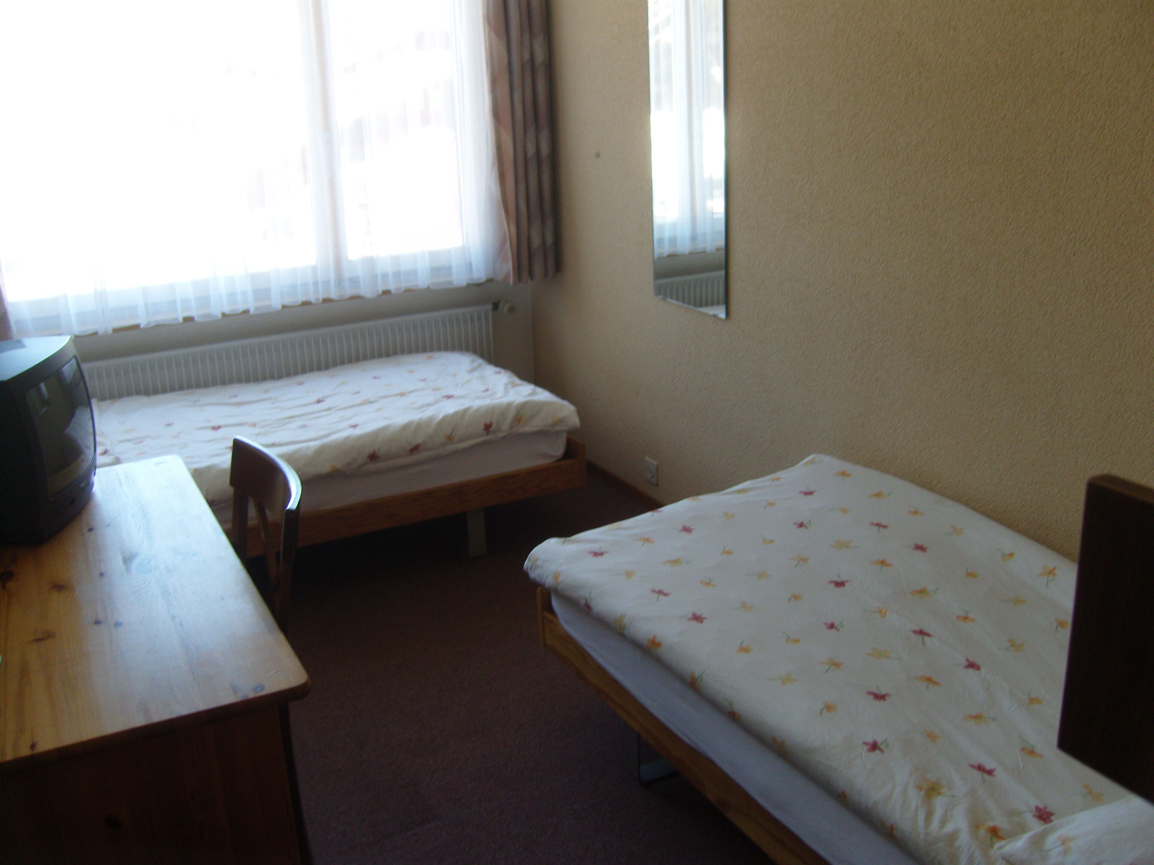 A small, simple room with two single beds, a wooden desk, and a chair.