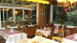 Un comedor con mesas dispuestas para una comida, rodeado de sillas y decorado con flores y plantas en macetas.