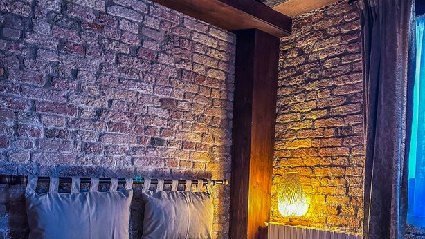 Une chambre avec un mur en briques, des poutres en bois, un lit avec des oreillers, une table de chevet et une lampe fixée au mur.