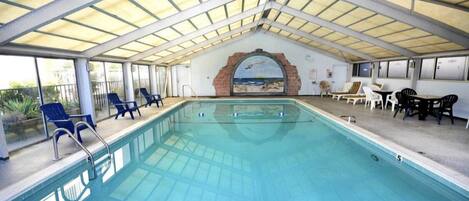 An indoor swimming pool with a glass ceiling, lounge chairs, and a mural of a beach scene.