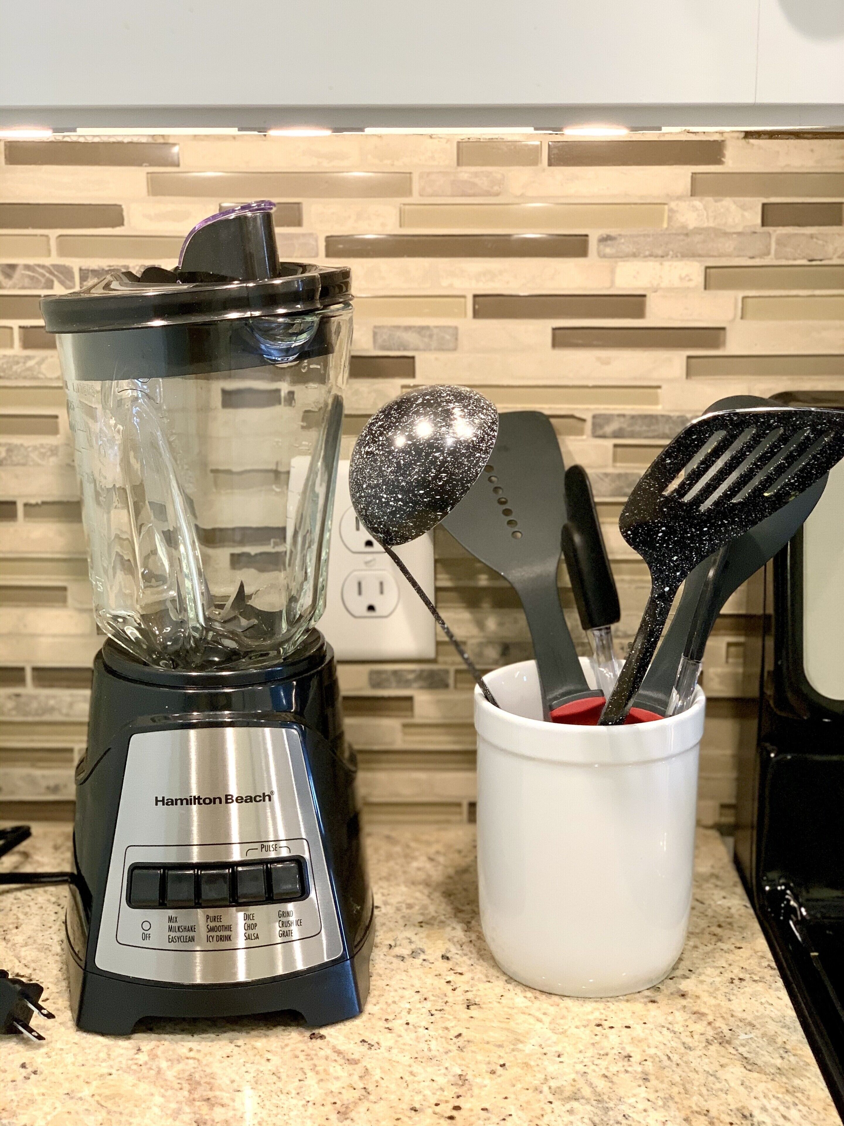 Eine Küche mit einem Hamilton Beach Mixer, einer weißen Tasse mit Besteck und einer gefliesten Spritzschutzwand.