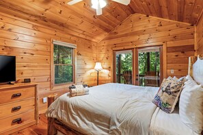 A wooden cabin bedroom with a bed, dresser, TV, and windows.
