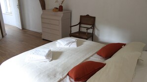 Een opgemaakt bed met witte lakens en twee rode kussens, een houten stoel en een witte dressoir in een kamer met houten vloer en witte muren.