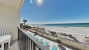 A balcony with a view of a beach, lounge chairs, and a palm tree. - 🌊 Kima 103 — Gulf Front 2BR Condo with Panoramic Views & Private Balcony (Madeira Beach)
