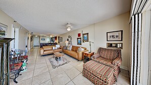 A spacious living room with a brown leather sofa, a patterned armchair, and a coffee table. A ceiling fan is visible, and the room has a tiled floor. - 🌊 Kima 103 — Gulf Front 2BR Condo with Panoramic Views & Private Balcony (Madeira Beach)