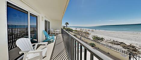 A balcony with a view of the beach, white chairs, and a railing.