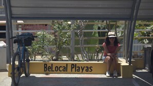 Une personne est assise sur un banc, à proximité duquel est garé un vélo. Le banc porte une pancarte sur laquelle on peut lire : « BeLocal Playas ».