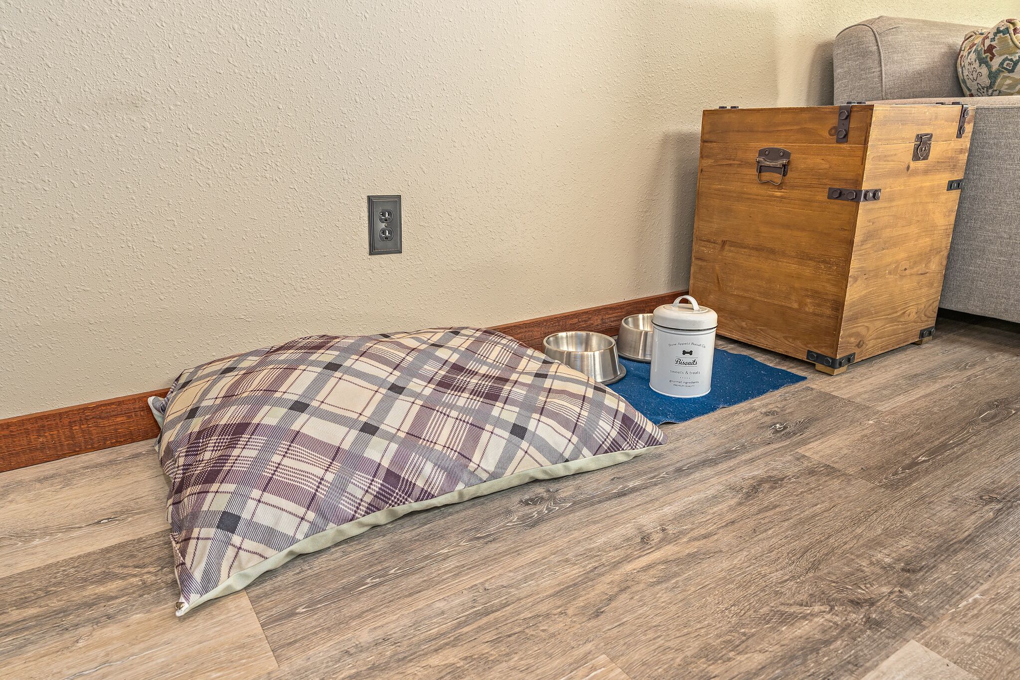A room with a wooden floor, a plaid pillow, a metal bowl, and a biscuit tin.