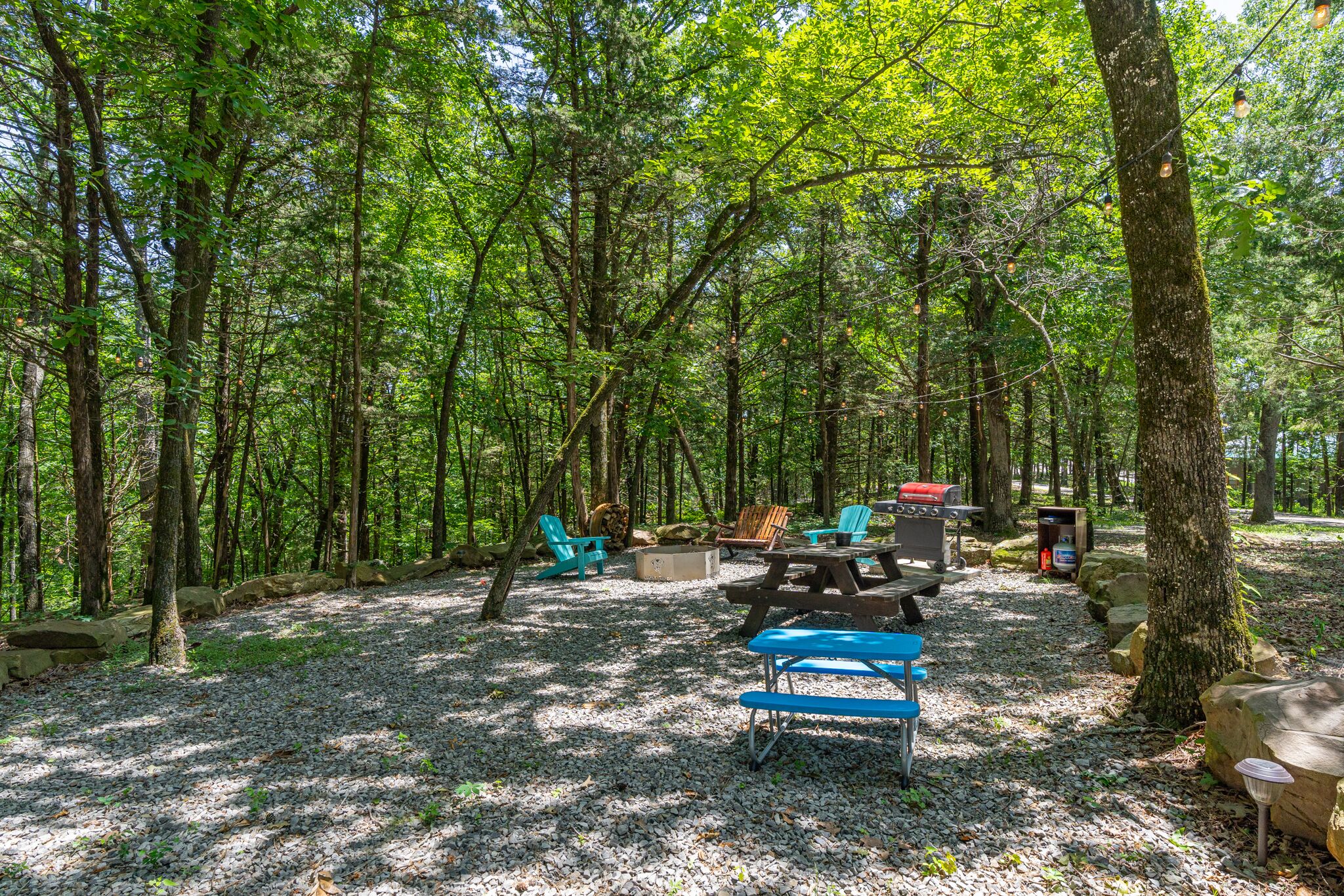Un terrain de camping situé dans une forêt, équipé de tables de pique-nique, de chaises et d’un barbecue.