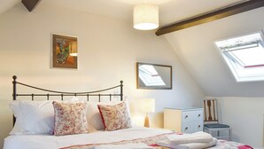 Une chambre à coucher avec un lit, des oreillers, une couette, une table de chevet avec une lampe, une commode, une chaise et une lucarne.