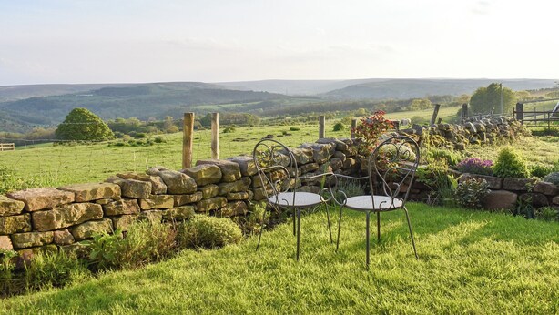 Un mur de pierres, avec une table et des chaises en métal, se trouve dans un champ verdoyant, offrant une vue sur des collines ondoyantes.