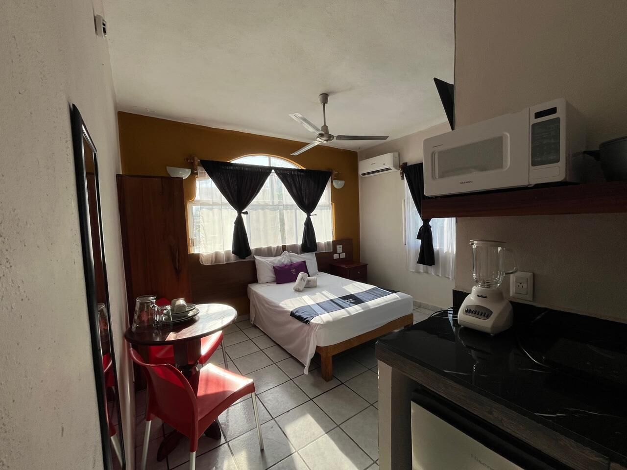 Ein kompaktes Hotelzimmer mit Bett, einem kleinen Esstisch, einem roten Sessel, einem Deckenventilator, einer Mikrowelle und einem Mixer.