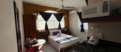 Ein kompaktes Hotelzimmer mit Bett, einem kleinen Esstisch, einem roten Sessel, einem Deckenventilator, einer Mikrowelle und einem Mixer.
