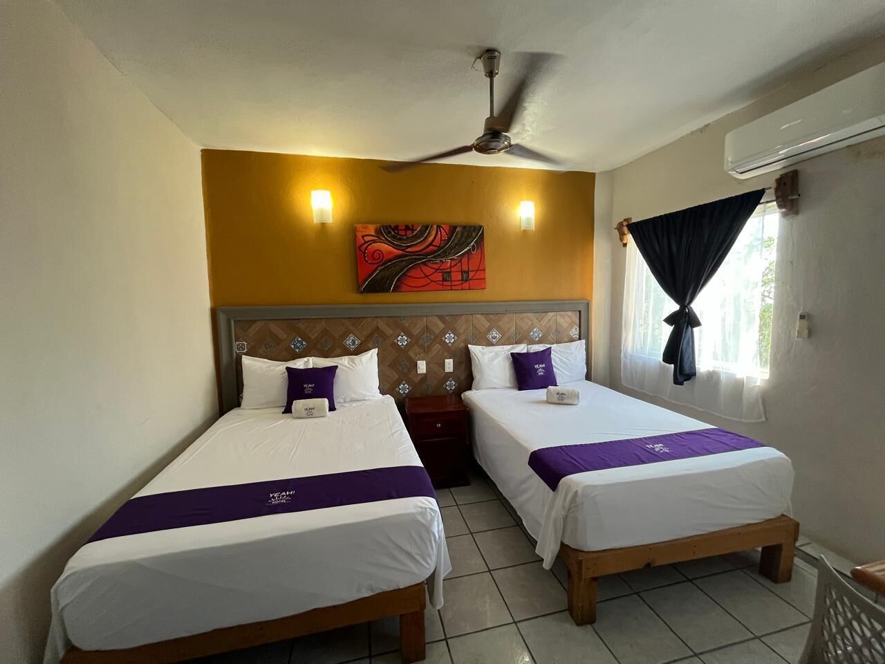 Una habitación de hotel con dos camas individuales, un ventilador de techo, un equipo de aire acondicionado, un ventanal con cortinas y un adorno en la pared.