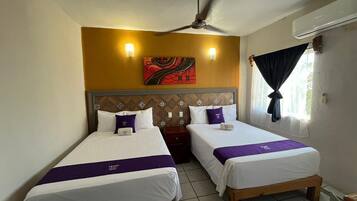 Una habitación de hotel con dos camas individuales, un ventilador de techo, un equipo de aire acondicionado, un ventanal con cortinas y un adorno en la pared.