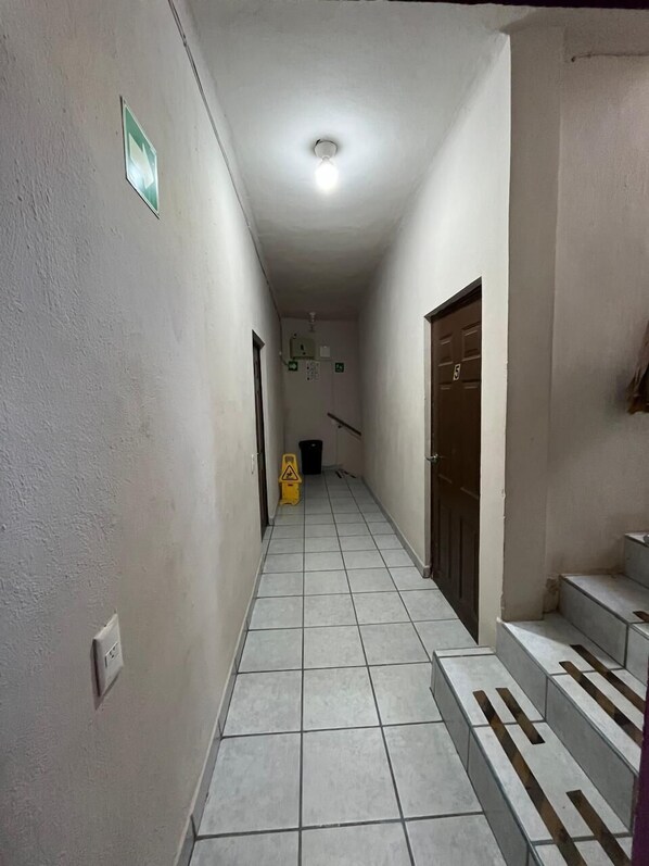 Un pasillo estrecho con suelo de baldosas, una lámpara en el techo, una puerta con número y un pequeño cubo de basura.