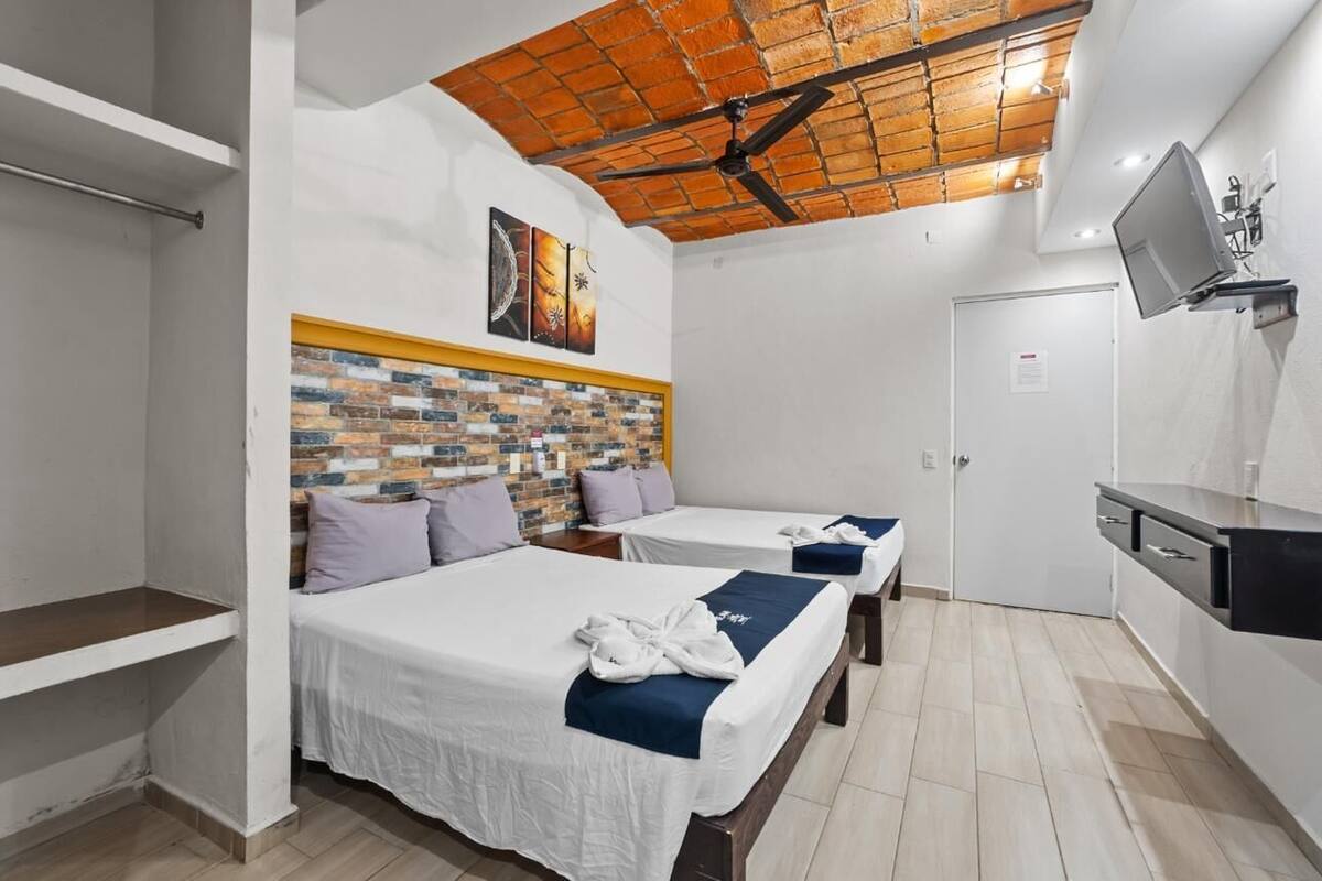 Habitación de hotel con dos camas, cabecero de ladrillo, ventilador de techo, televisor montado en la pared y una pequeña kitchenette.