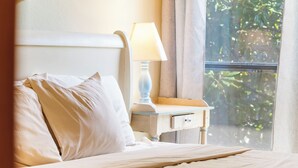 A neatly made bed with white linens and pillows, a bedside table with a lamp, and a window with a view of greenery.
