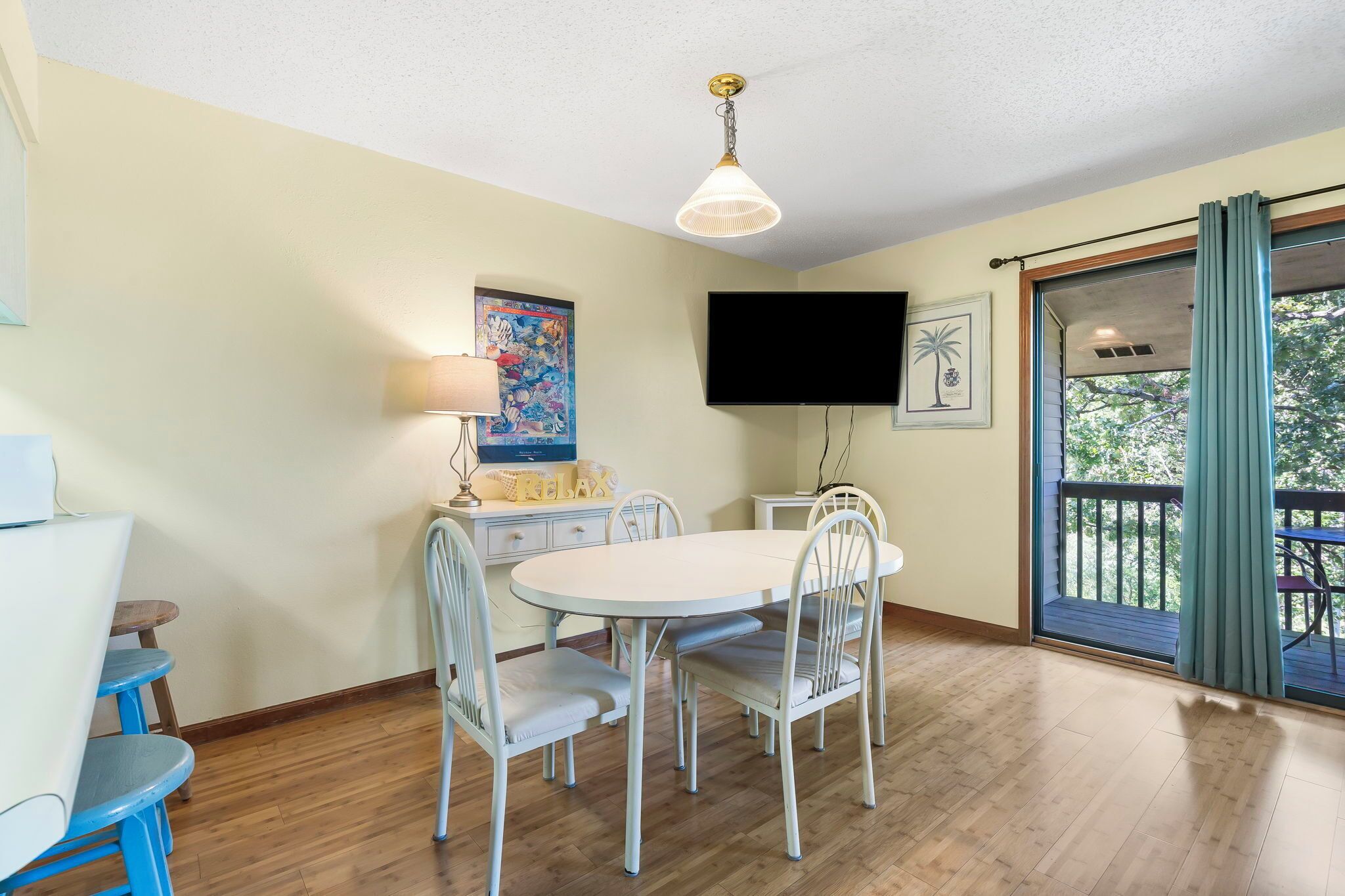 A dining area with a white table and chairs, a ceiling fan, a flat-screen TV, and a window with a view of greenery.