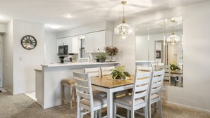 Un coin repas avec une table en bois et des chaises blanches, une cuisine en arrière-plan et un grand miroir au mur.