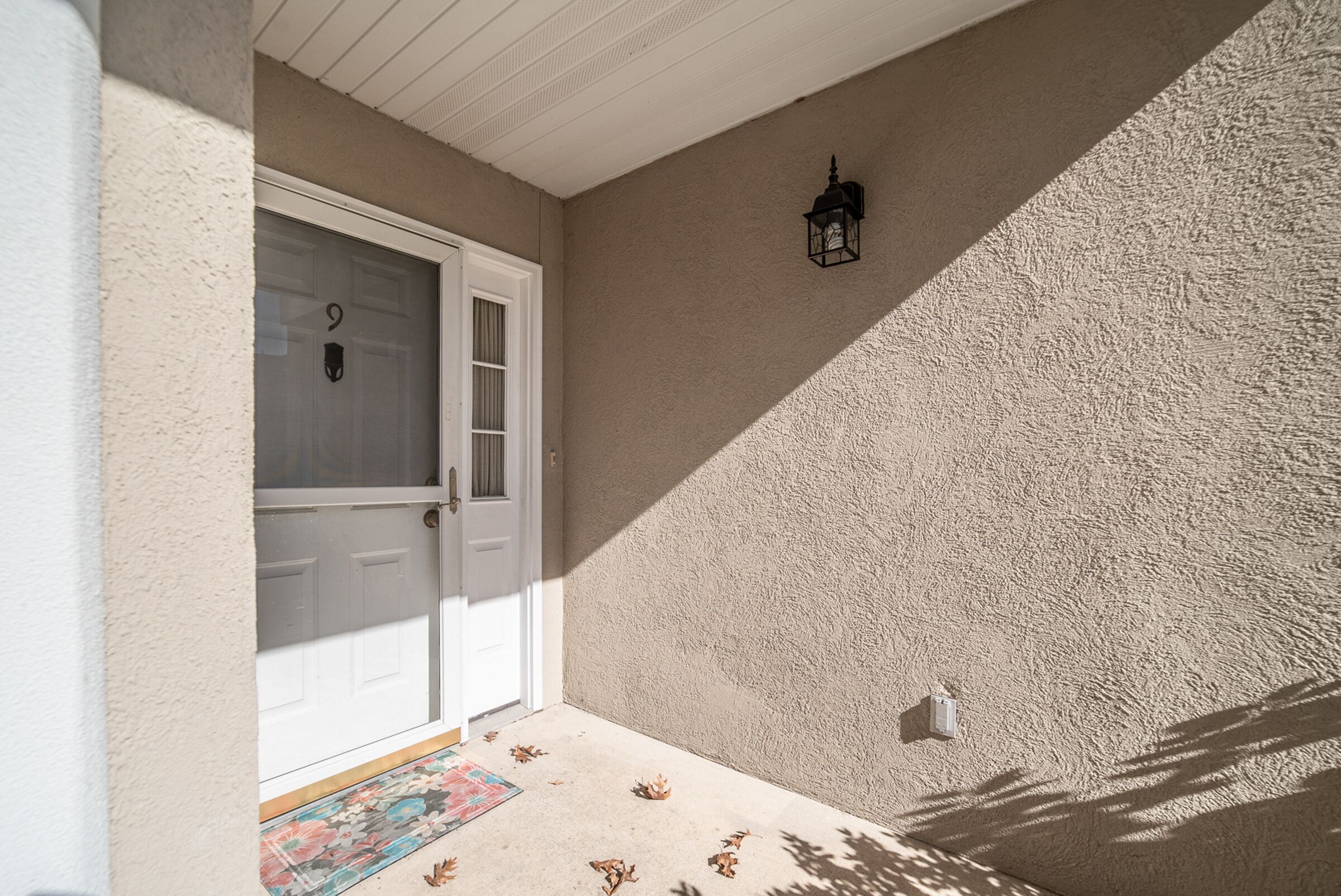 A white door with a glass panel, a textured beige wall, a small black lantern, and a colorful mat at the entrance.