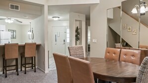 Un coin repas avec une grande table en bois et six chaises en cuir marron. À gauche, on trouve un espace cuisine avec un réfrigérateur et des tabourets de bar. Un grand miroir reflète une partie de la pièce, et un escalier est visible au fond.