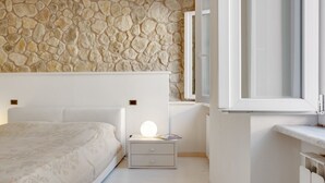 Un dormitorio con pared de piedra, cama con colcha estampada, mesita de noche con lámpara y ventana con vistas.