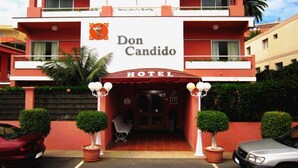 Un hotel llamado Don Candido con fachada roja y blanca, una entrada central y plantas en macetas.