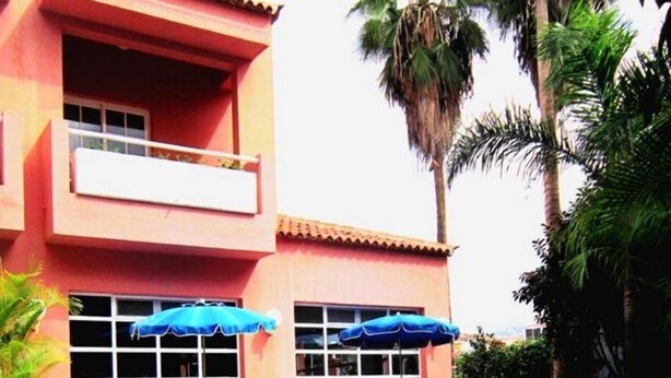 Un área de piscina con sillas de descanso y sombrillas, un edificio de fachada roja y palmeras.