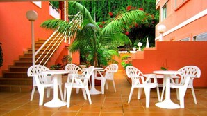 Un patio con sillas y mesas de plástico blanco, rodeado de paredes anaranjadas y una escalera.