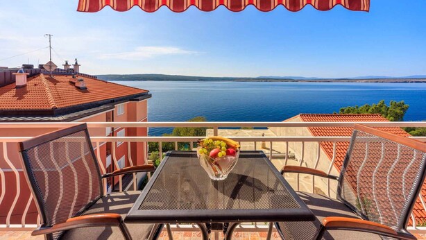 Een balkon met een tafel, stoelen, een schaal fruit en uitzicht op zee.