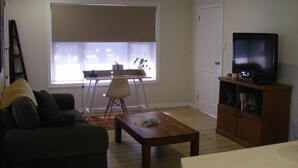 A living room with a dark-colored sofa, a wooden coffee table, a television on a wooden stand, and a window with blinds.