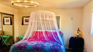 A bedroom with a bed covered in a colorful blanket, a canopy, a chandelier, a small table with a lamp, and a green chair.