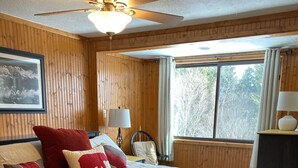 Ein Schlafzimmer mit einem großen Bett, einem Deckenventilator, einem Fenster mit Vorhängen, einem Sessel und einem Nachttisch mit Lampe.