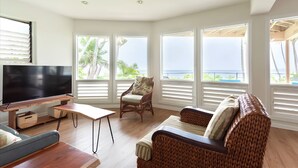 A spacious living room with a large flat-screen TV, wooden furniture, and large windows offering a view of the ocean. - OCEANFRONT DUPLEX, with POOL, SAUNA and steps from BEACH (Hilo)