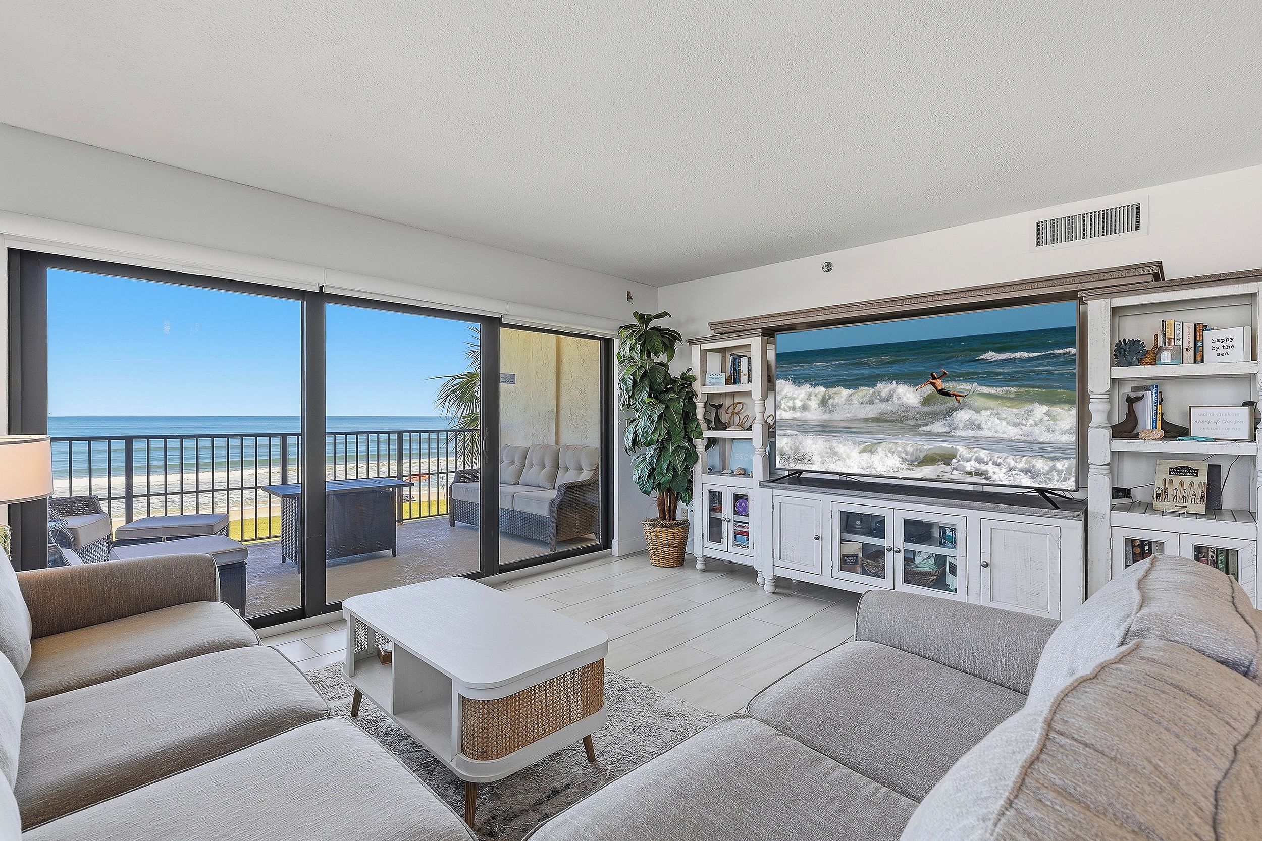 A living room with a large TV displaying a beach scene, a balcony with a view of the ocean, and a cozy seating area with a coffee table.