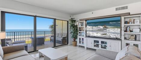 A living room with a large TV displaying a beach scene, a balcony with a view of the ocean, and a cozy seating area with a coffee table.