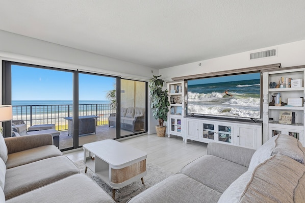 A living room with a large TV displaying a beach scene, a balcony with a view of the ocean, and a cozy seating area with a coffee table.