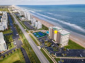 Vista aérea de un hotel costero con un amplio estacionamiento, cancha de tenis y vistas a la playa.